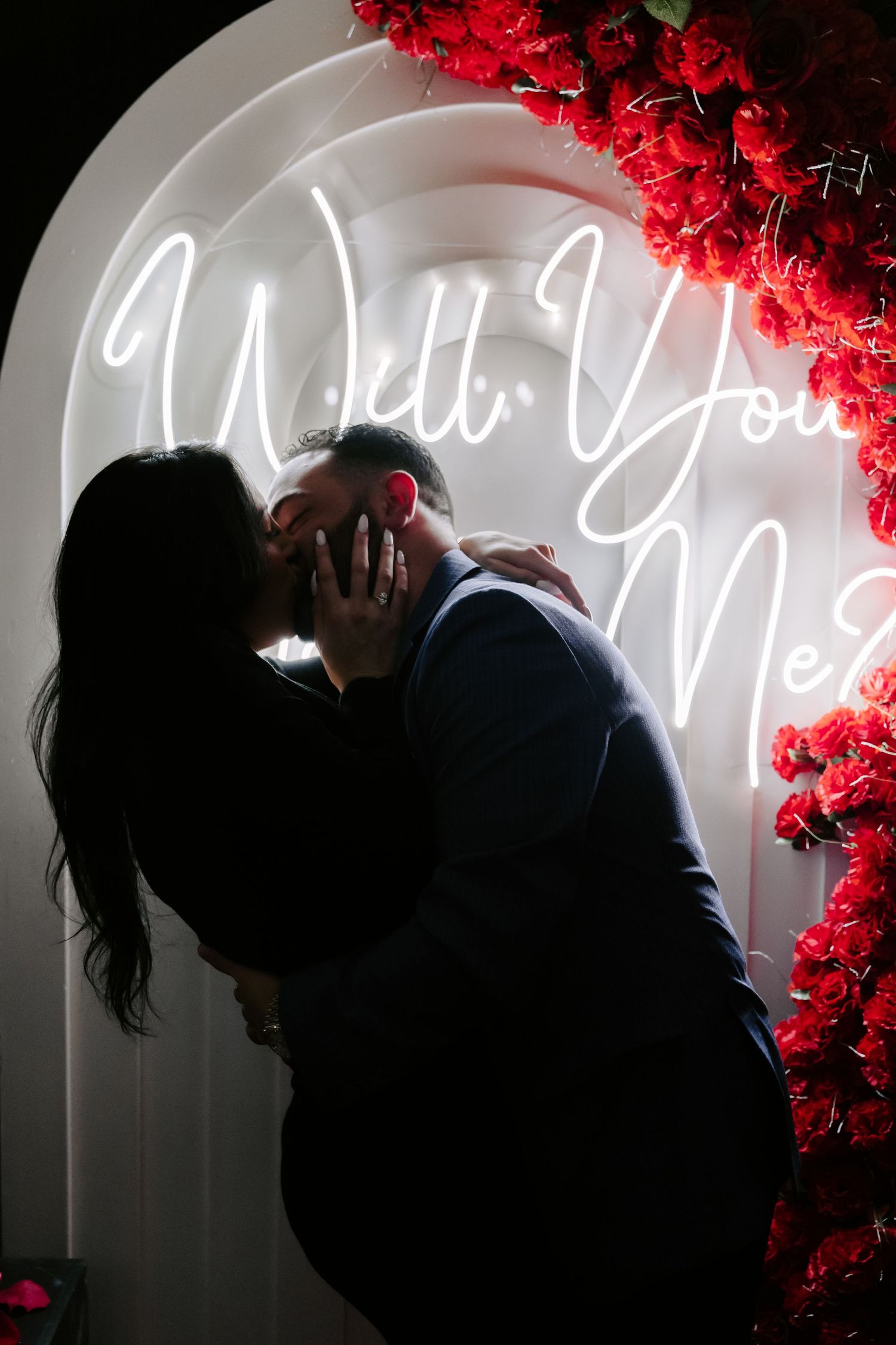 A silhouetted couple embraces under a neon 'Will You Marry Me' sign with red roses.