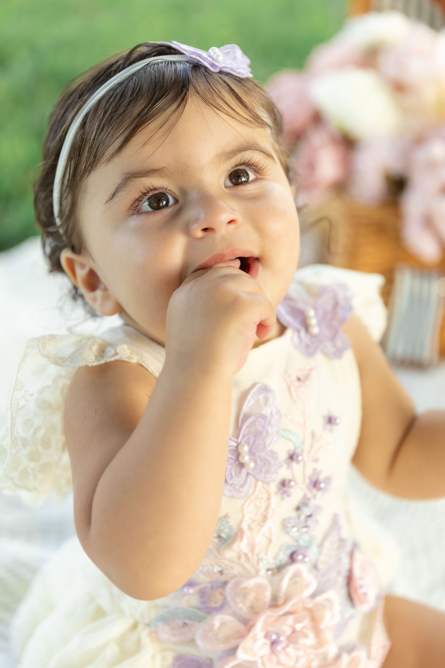 A small child in a white and purple dress with a headband looks upward while touching their face during a special occasion.