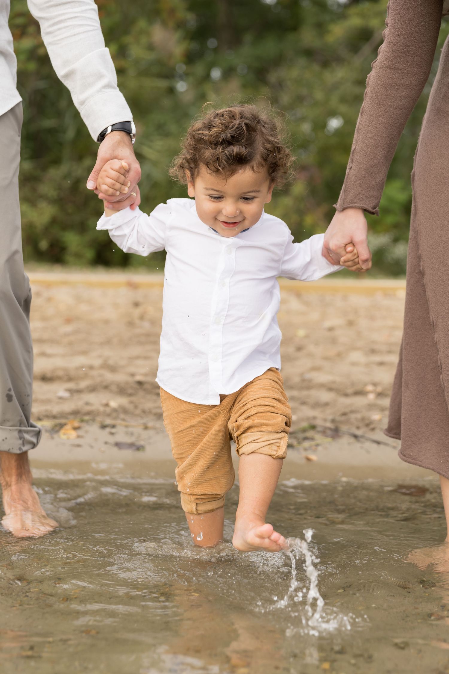 Small child splashes in shallow water while holding hands with parents on beach wearing white shirt and khaki pants.