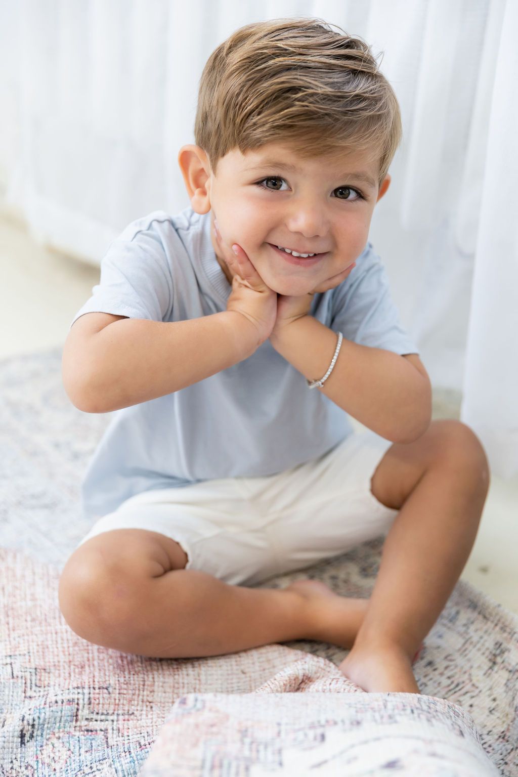 Smiling child in light blue shirt and shorts sitting cross-legged on blanket while resting head on hands.
