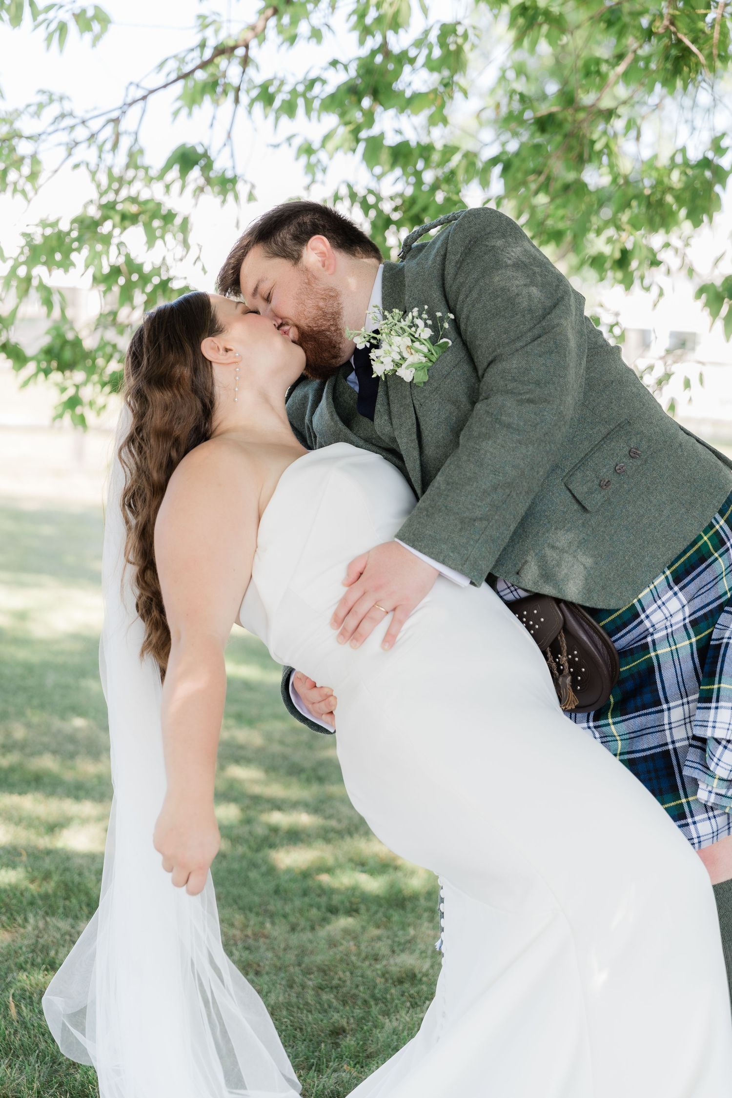 Groom in Scottish attire dips bride for a romantic kiss under green foliage.