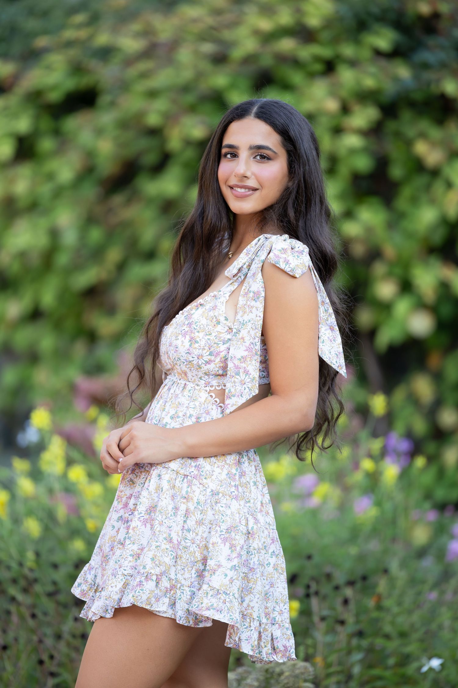 Woman in floral summer dress poses in garden setting with blooming flowers.