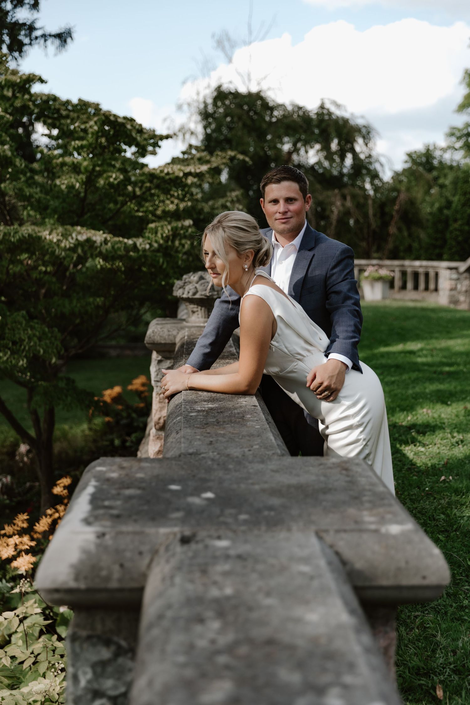 Couple in wedding attire posing by stone wall in sunlit garden setting.