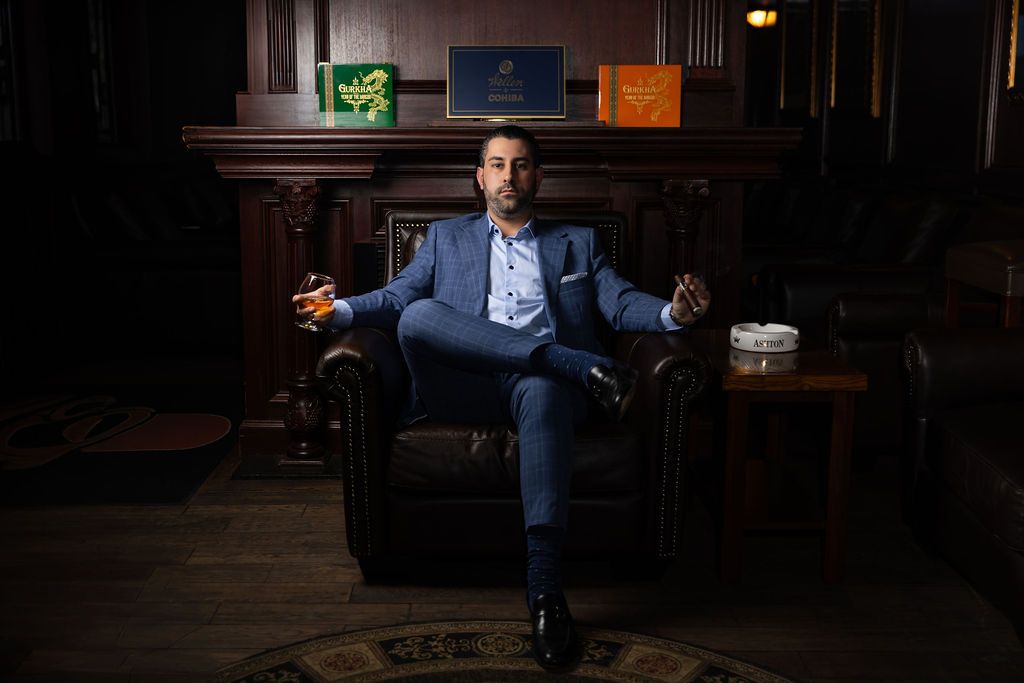 A well-dressed person sits confidently in a leather armchair at a dimly lit bar, holding a cocktail glass.