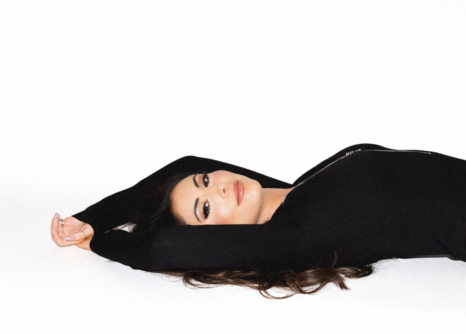 A person with long dark hair wearing a black outfit lies gracefully on a white background.