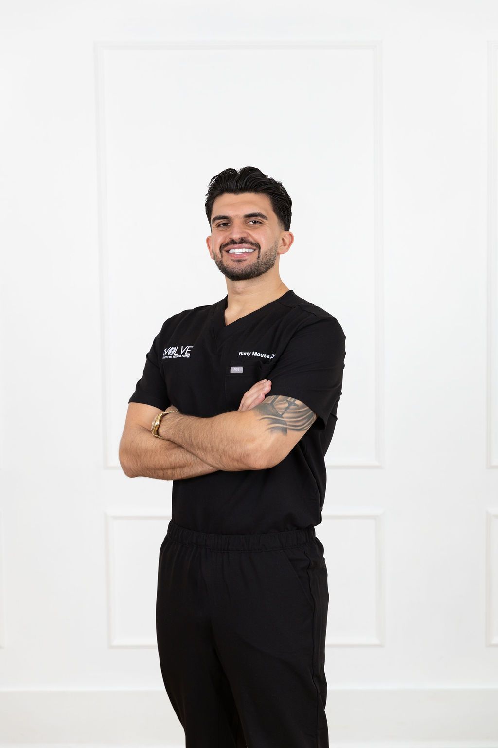 Healthcare professional in black scrubs stands with arms crossed and friendly smile against white background.