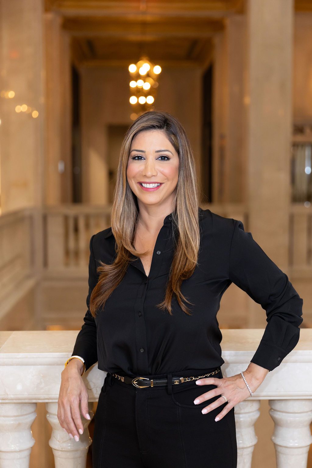 Stylish professional in all-black attire poses elegantly against a marble railing in an ornate interior.