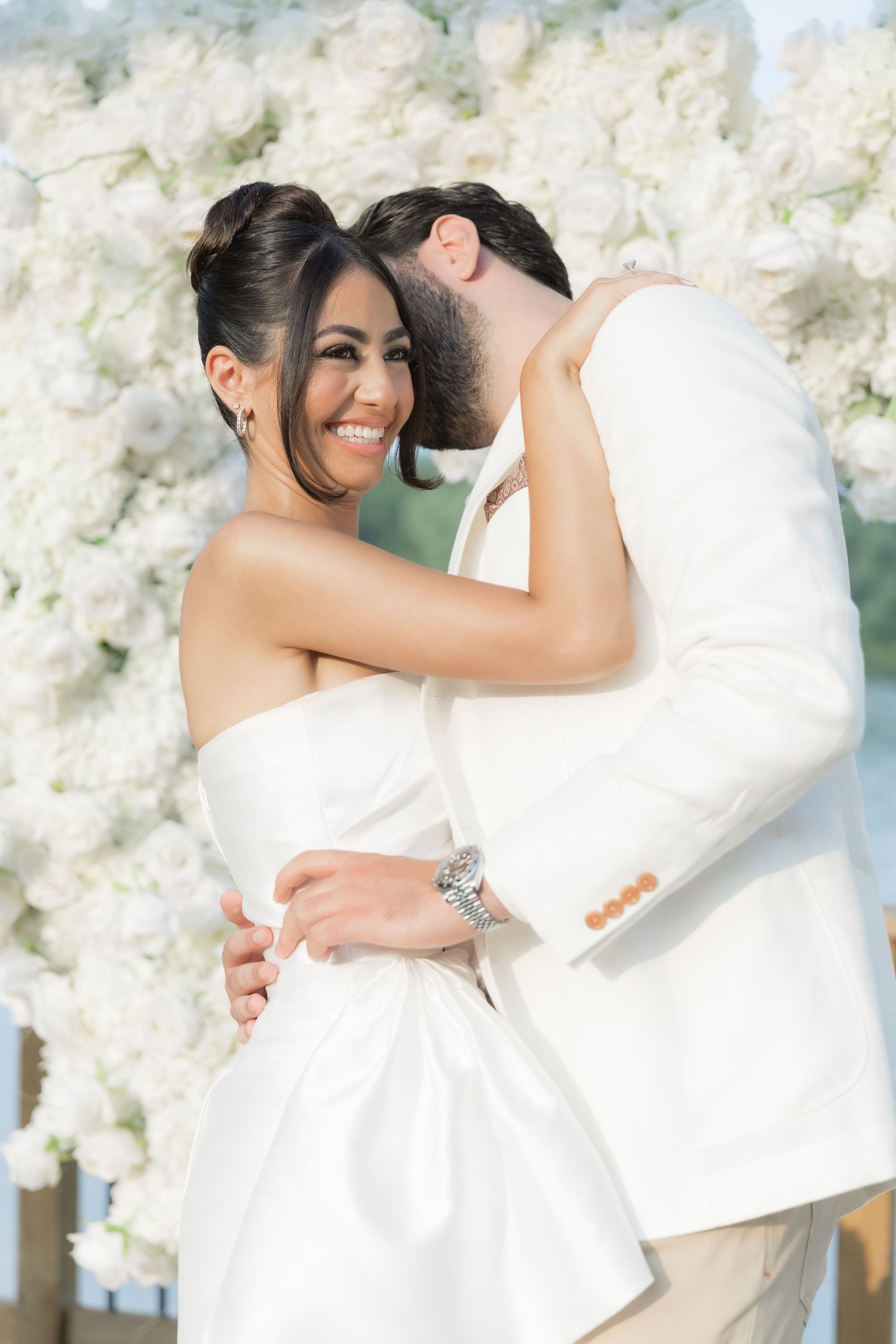 Joyful couple embraces in front of a white floral wall during their wedding celebration.