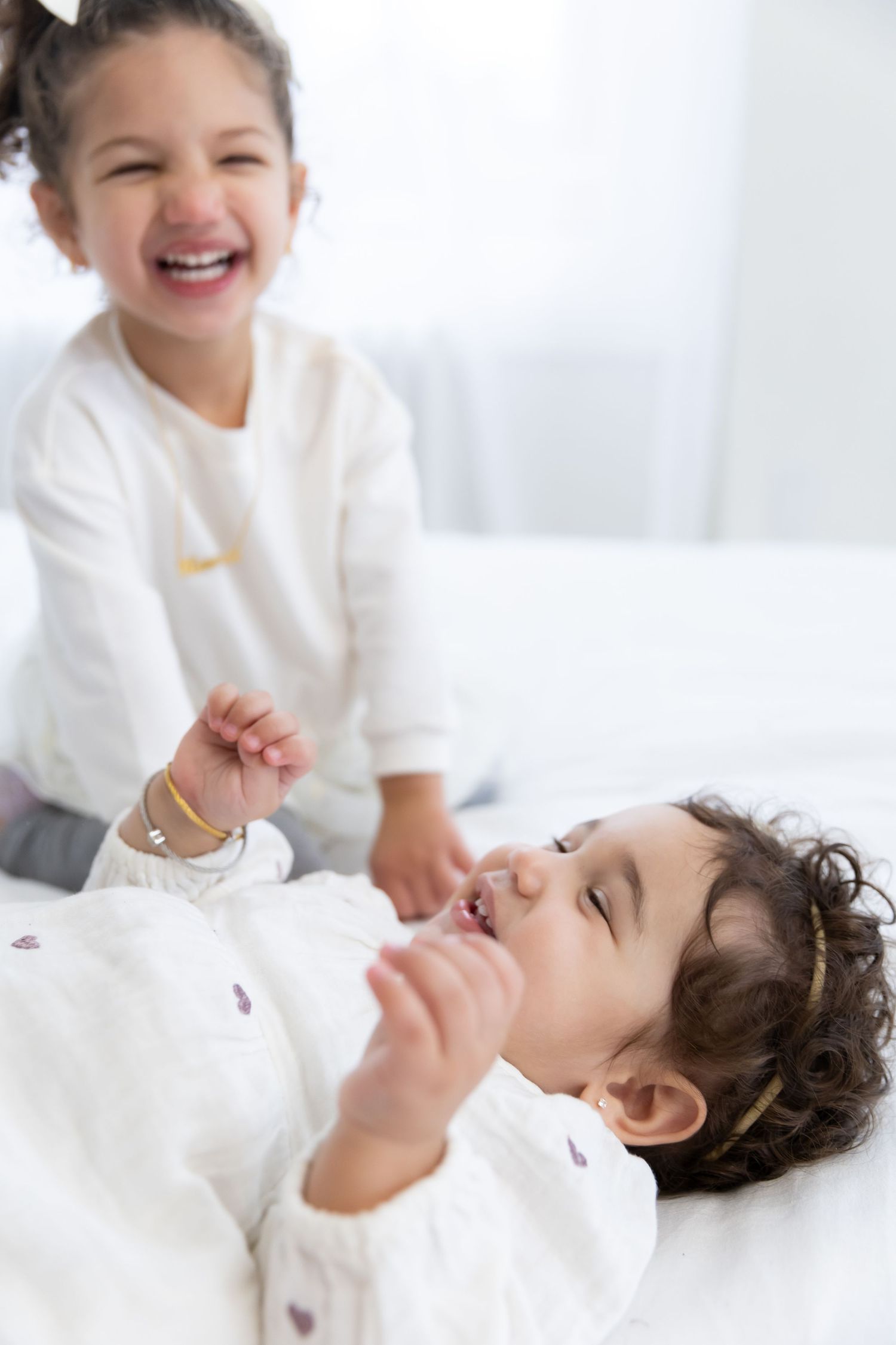 Two children in white clothes playing and laughing together on a bright bed.