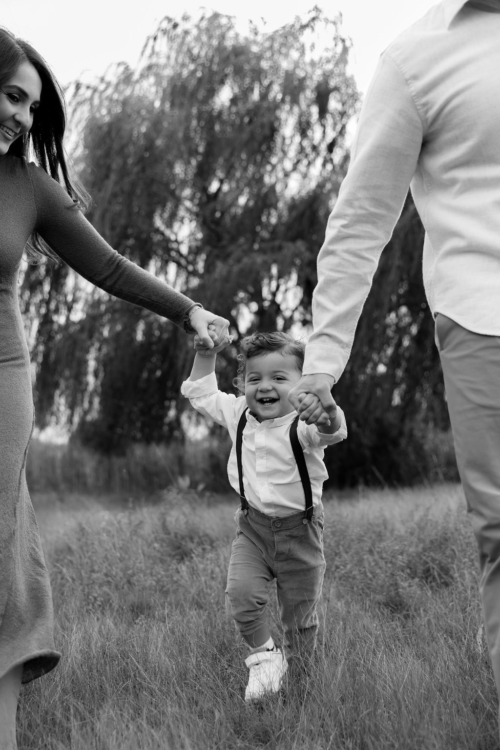 A joyful toddler in suspenders and white shirt walks between two adults holding hands in a grassy outdoor setting.