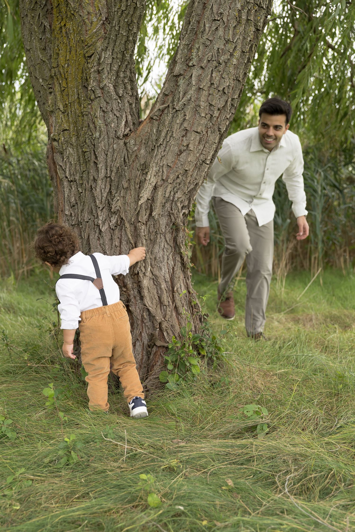 A playful scene around a large tree trunk in a grassy outdoor setting with two people dressed in light colored clothing.