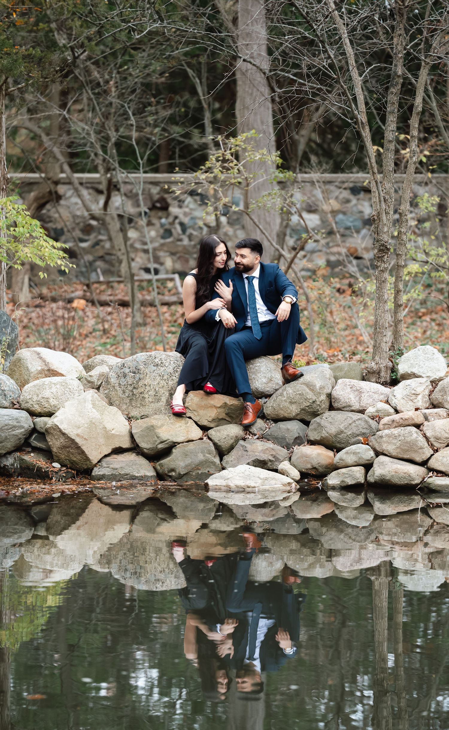 A romantic couple sitting on stone wall with their reflection in a serene pond below.