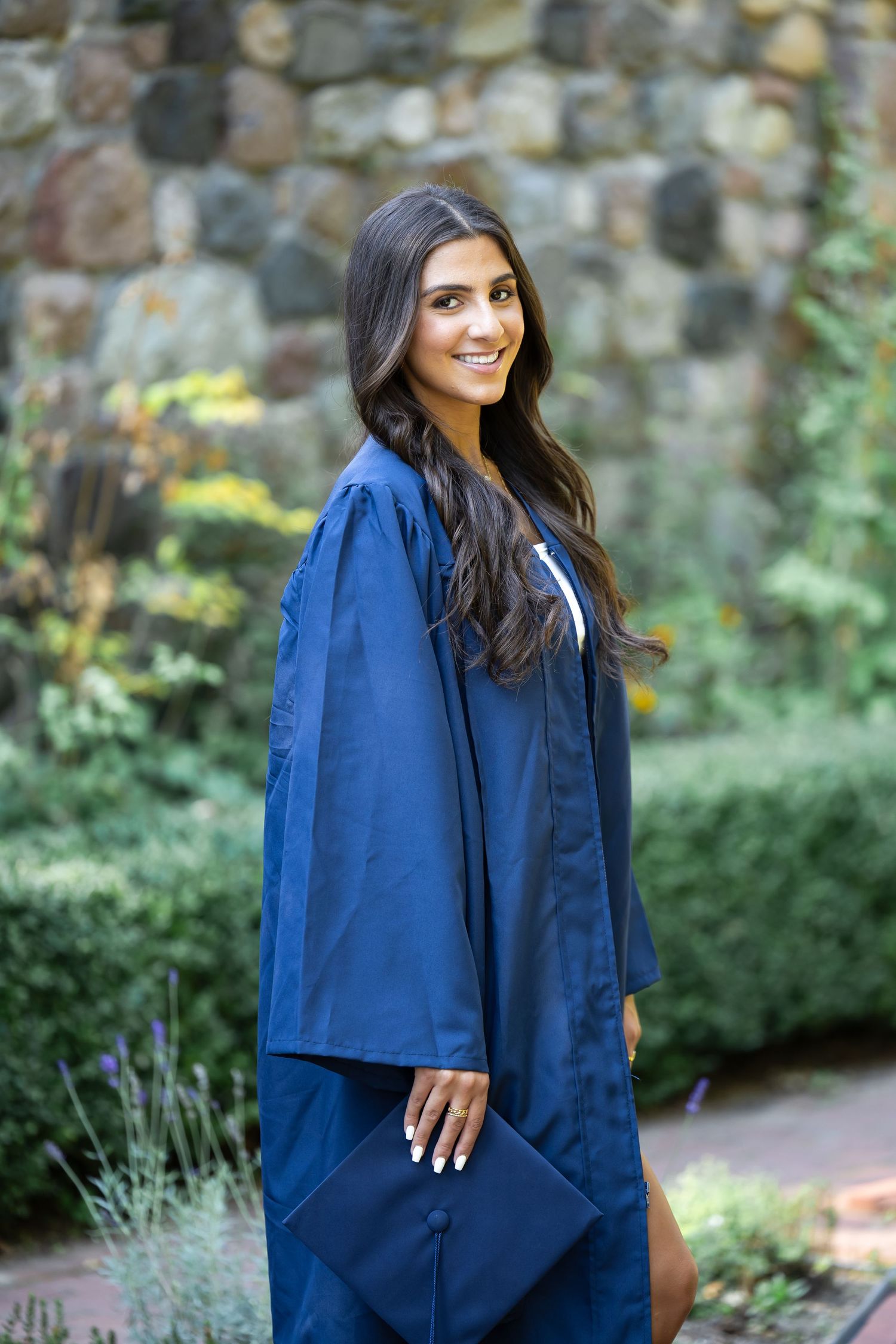 Graduate in blue cap and gown poses in garden setting with stone wall background.