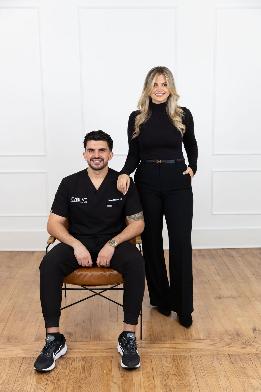 Professional portrait of two people in black outfits posing on hardwood floor against white wall with panel details.