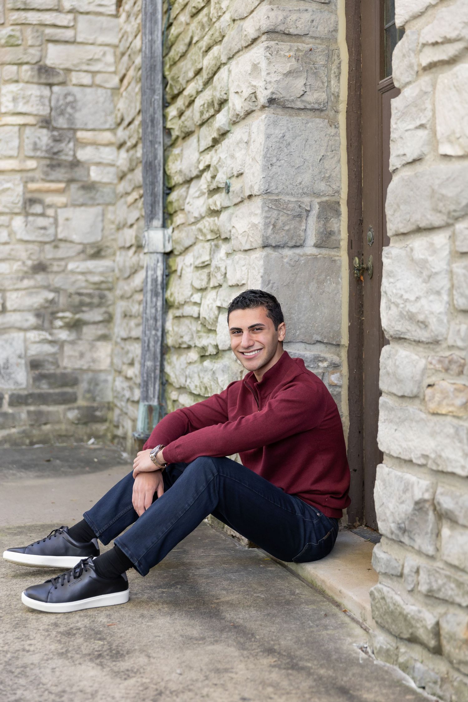 Man in burgundy sweater sitting against stone wall outdoors.