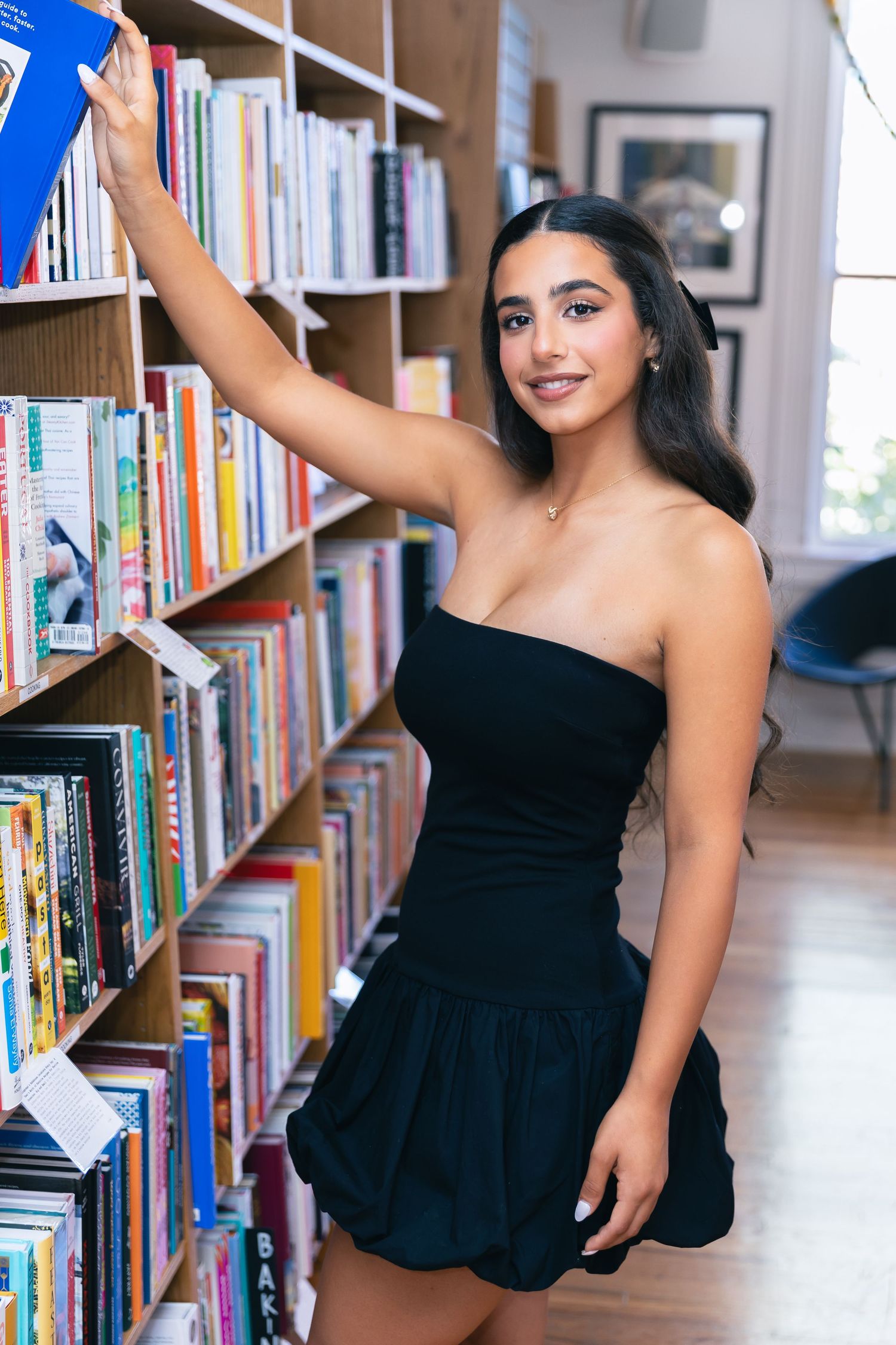 Person in black dress reaching for book on library shelf.