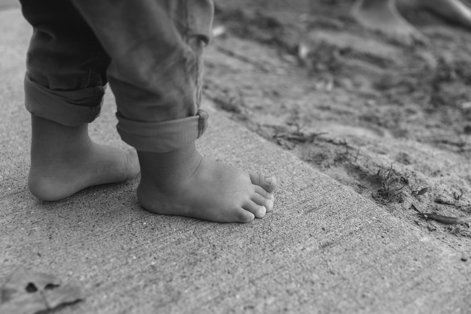 Black and white close-up photograph of bare feet standing on sand or textured ground.