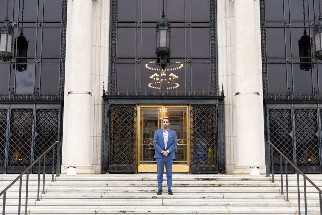 Someone stands on the steps of a stately building entrance with ornate black doors and white columns.