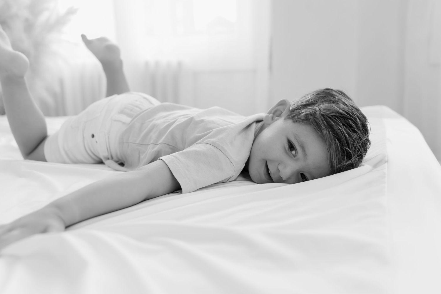 Black and white photo of child laying on white bedding in a contemplative pose with natural lighting.