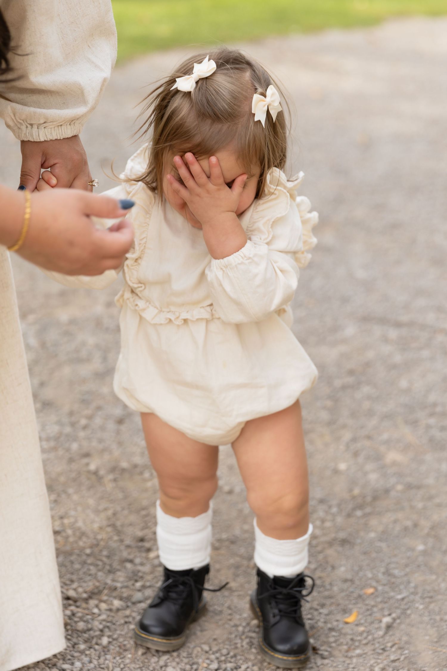 Small child in a white ruffled dress and combat boots covers face while standing on gravel path.