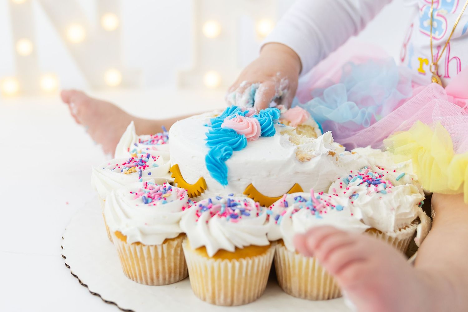 Hand decorating vanilla cupcakes with colorful sprinkles and blue frosting on a white serving plate.