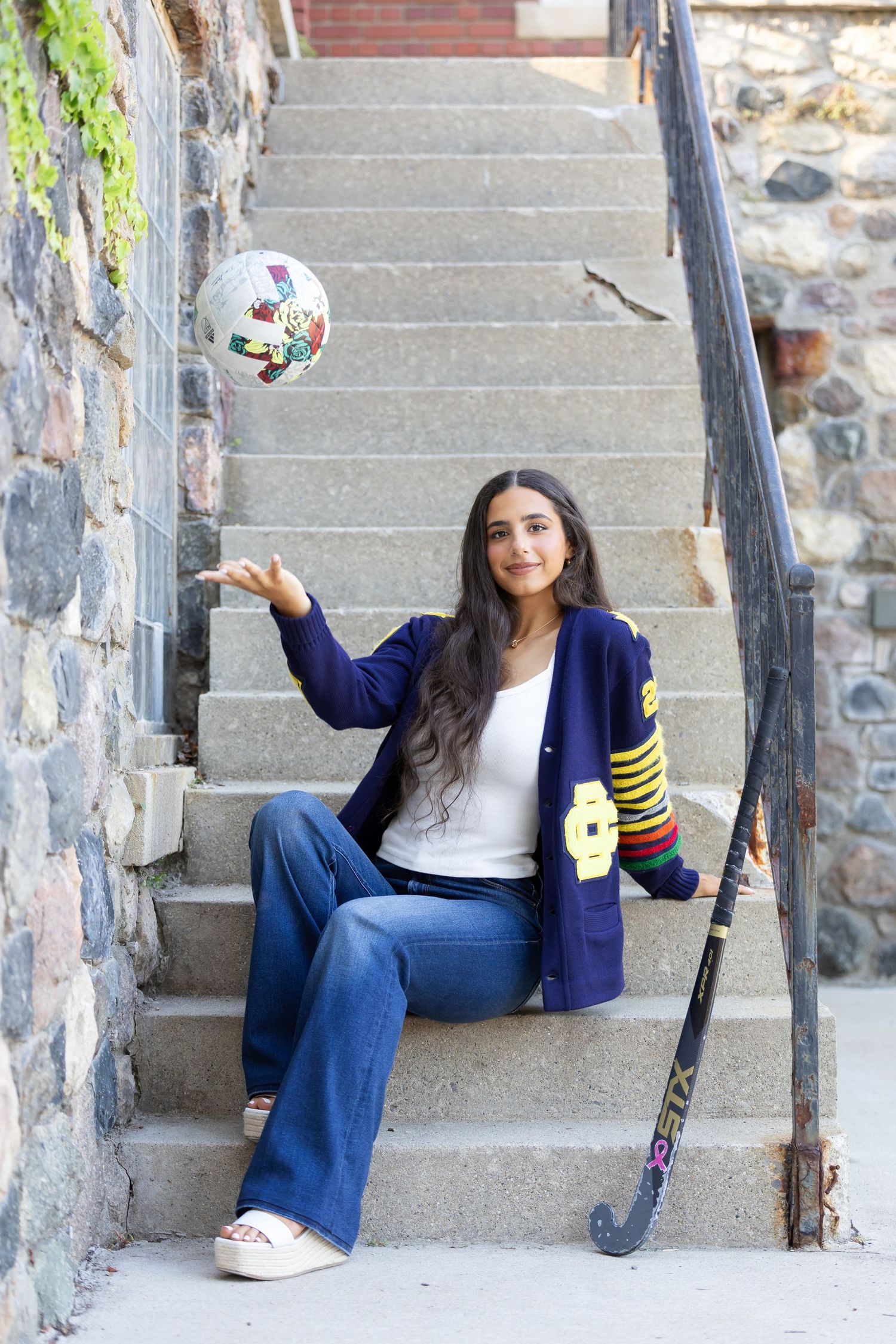 Person in varsity jacket sits on stone steps with field hockey stick and disco ball.