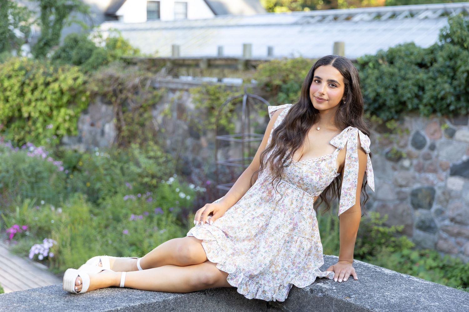 Woman in floral dress sitting on stone wall in garden setting.