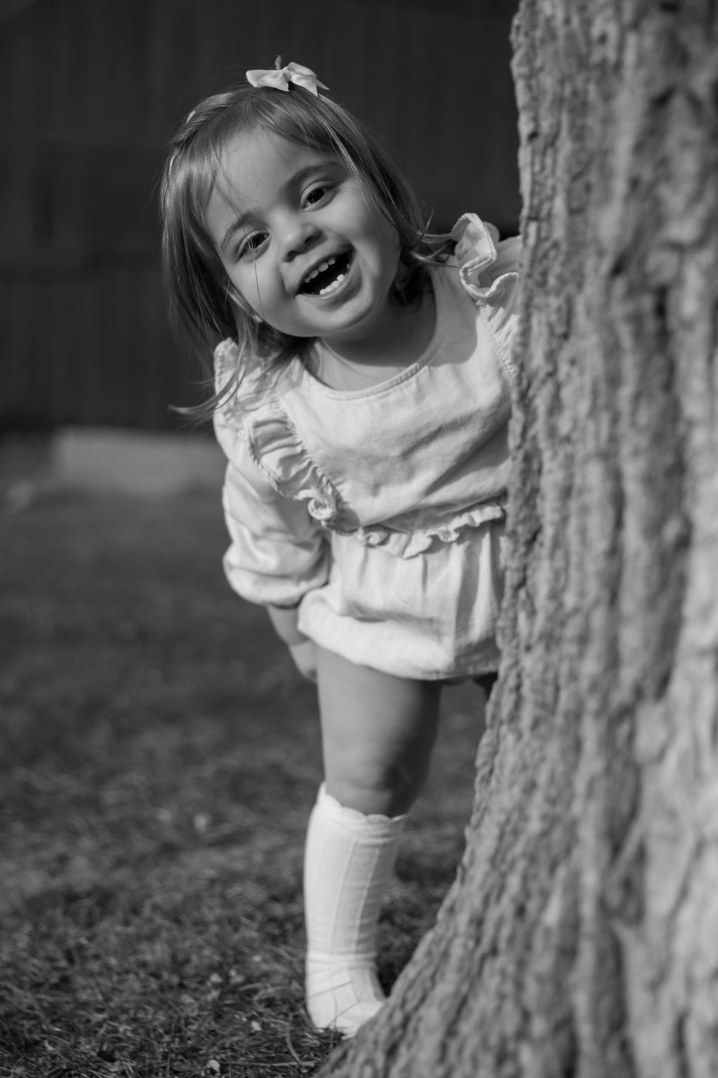 A joyful toddler in a white dress and knee socks peeks from behind a tree trunk in this black and white portrait.