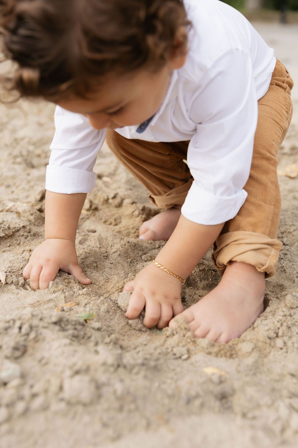 Small child in white shirt and brown pants explores and plays in sandy ground with bare feet.