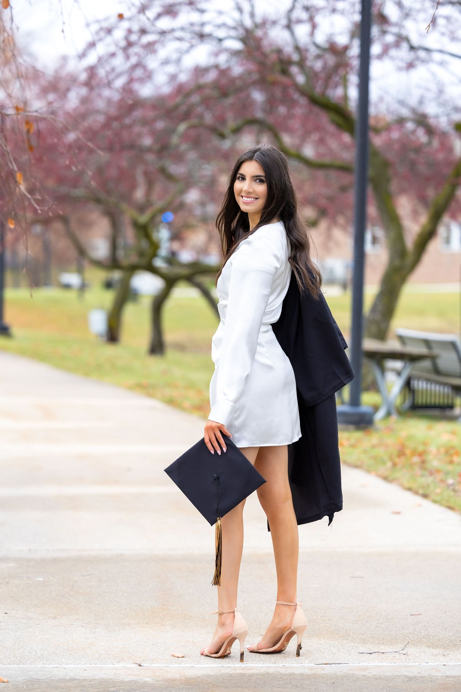 A woman in a white dress holds a graduation cap while posing outdoors near blooming trees.