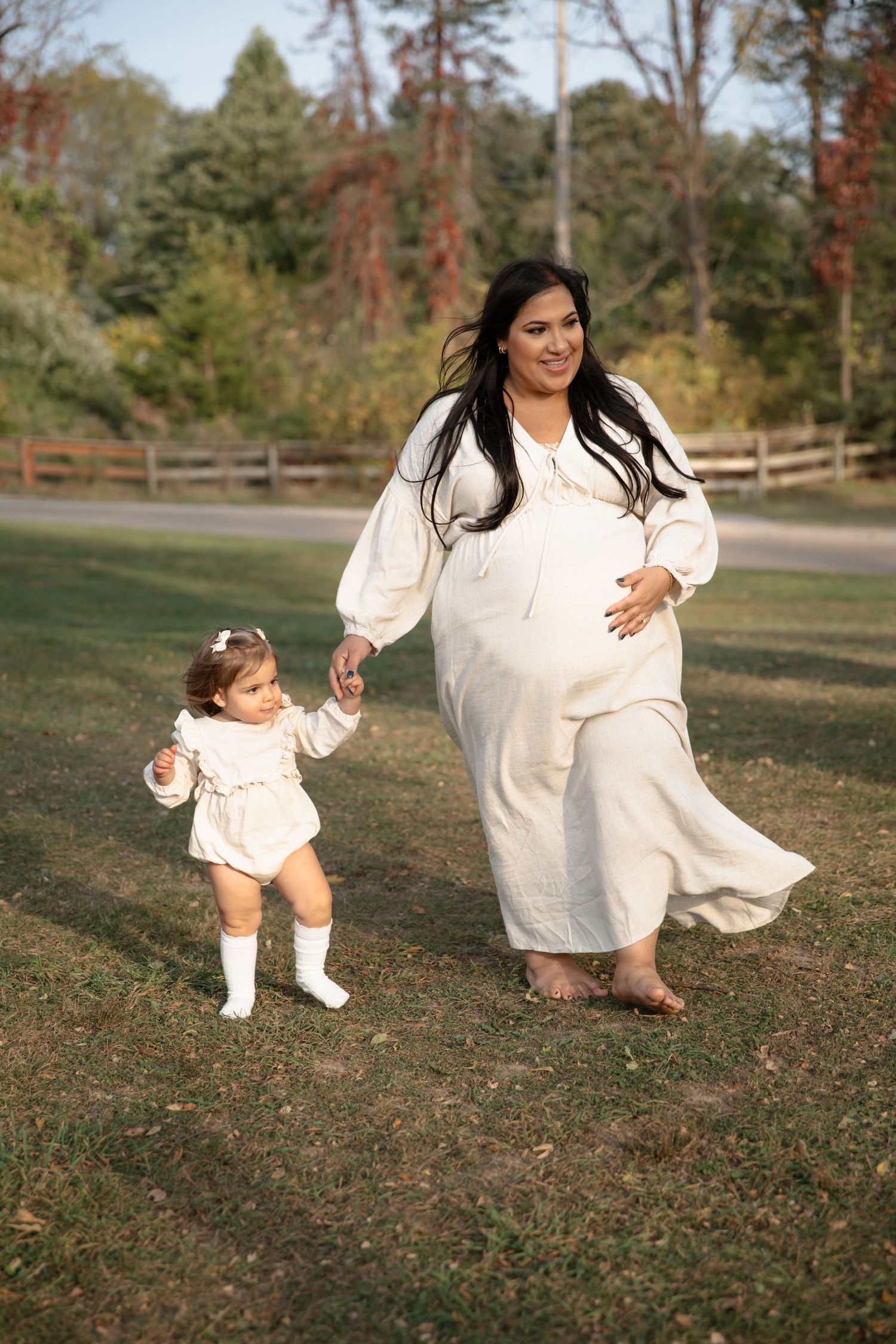 Mother and toddler in matching white dresses hold hands while walking in a park during autumn.