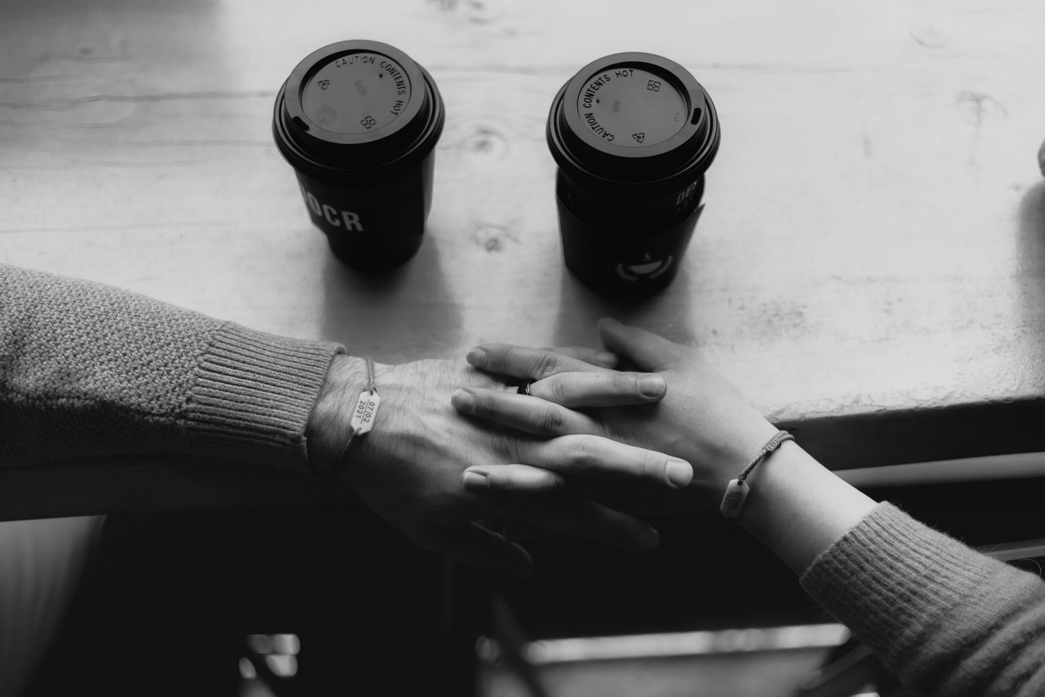 Close-up of intertwined hands next to two black coffee cups on a light surface.