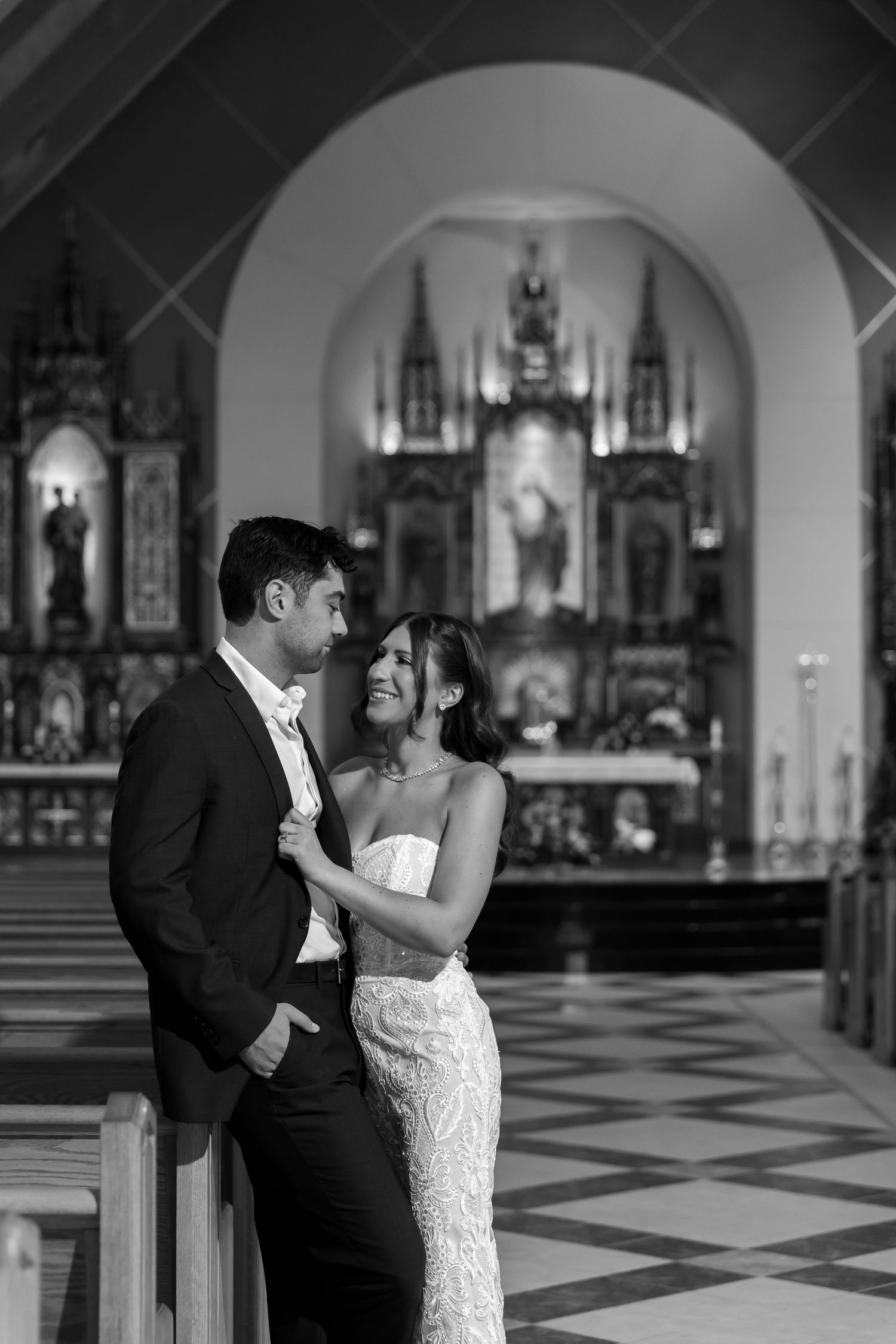 A couple shares an intimate moment in formal attire inside an ornate Catholic church with altar visible.