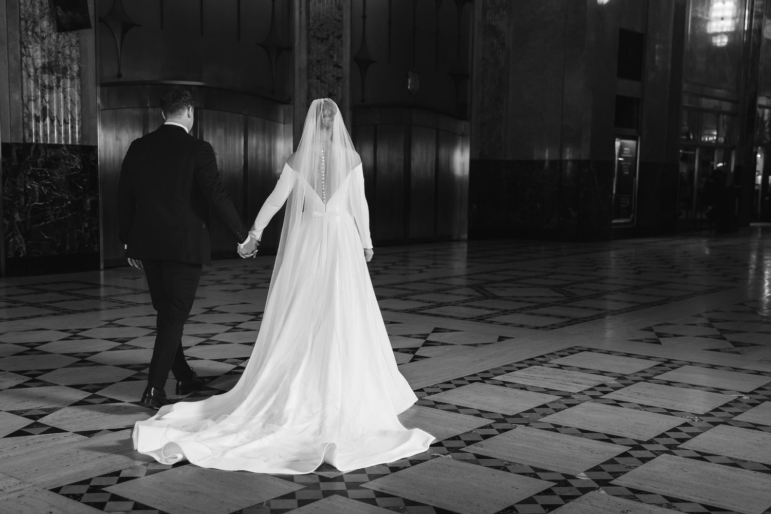Bride in flowing white dress walks with groom across patterned floor in dramatic black and white shot.