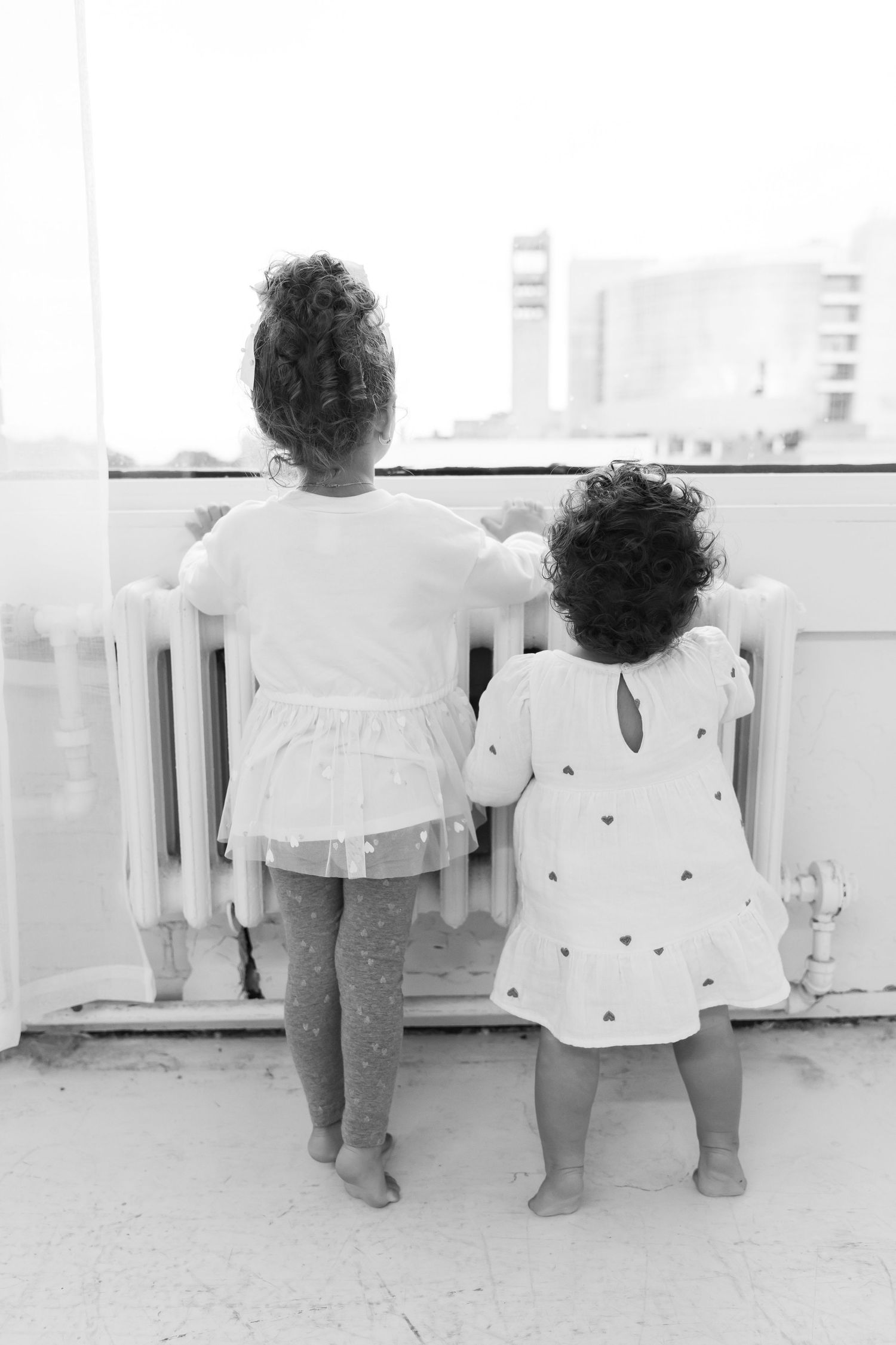 Two small children in white dresses stand at a railing overlooking a cityscape in black and white photography.