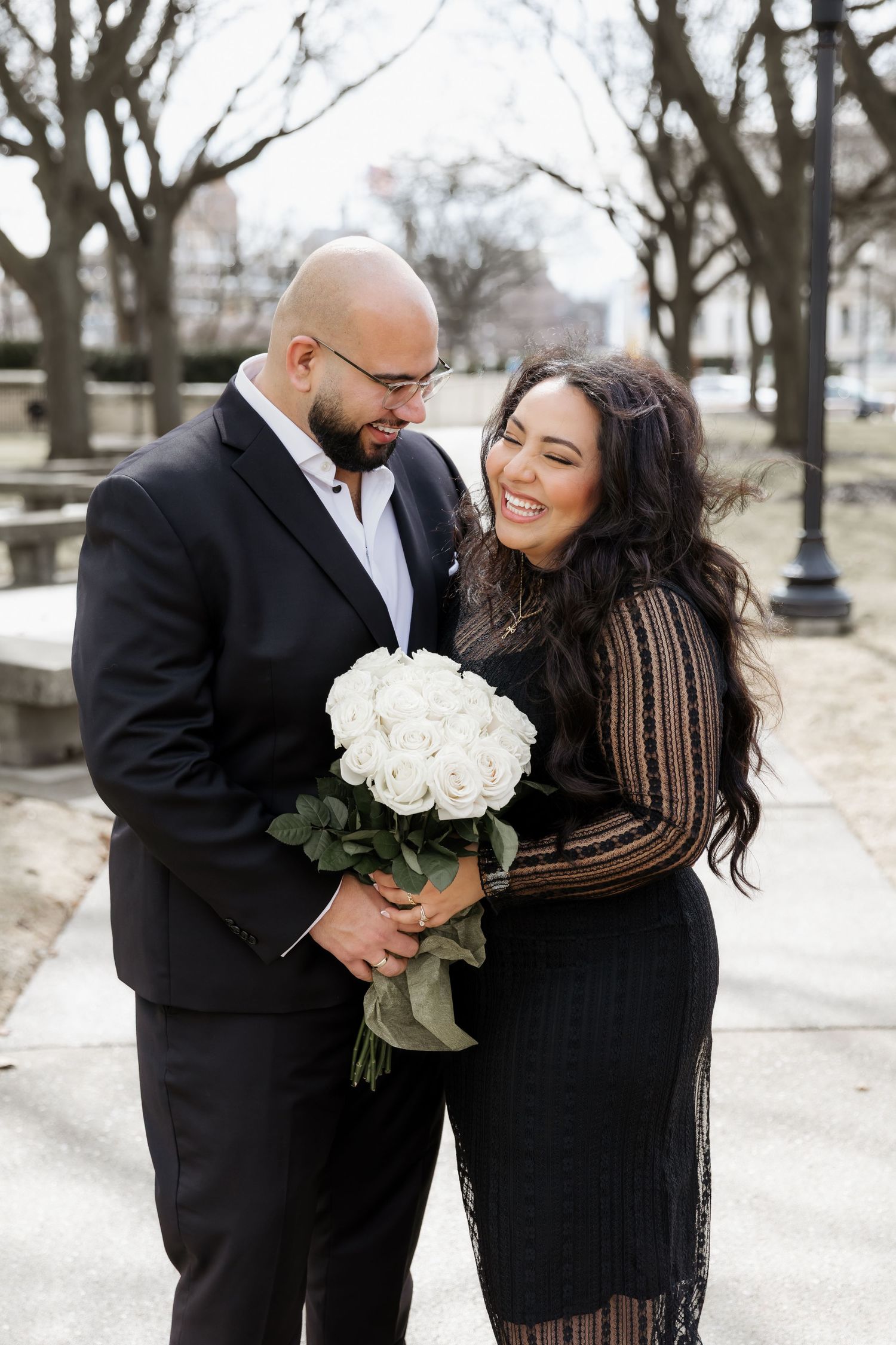 A couple shares a joyful moment while holding white roses against a winter backdrop.