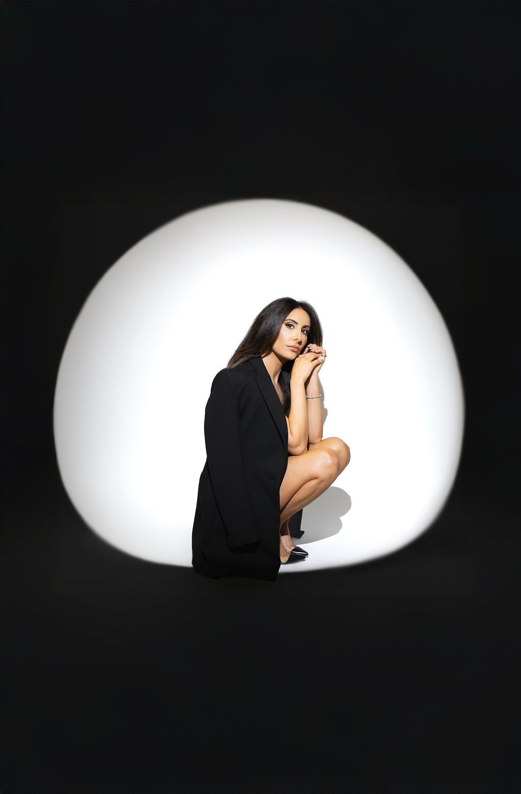 Artistic portrait of a figure in black crouching against a white circular backdrop on dark background.