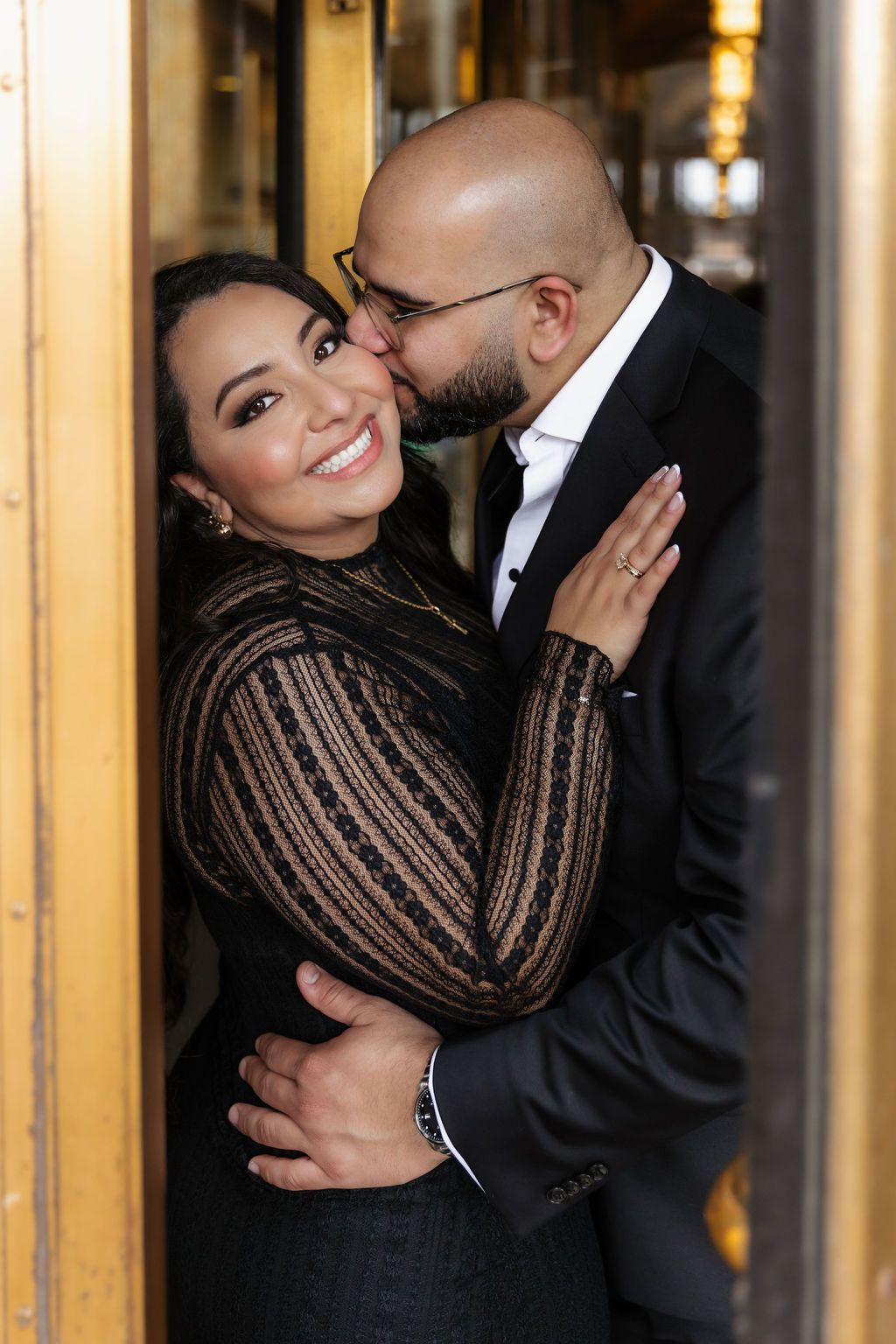 A romantic couple embraces in formal attire near a doorway.