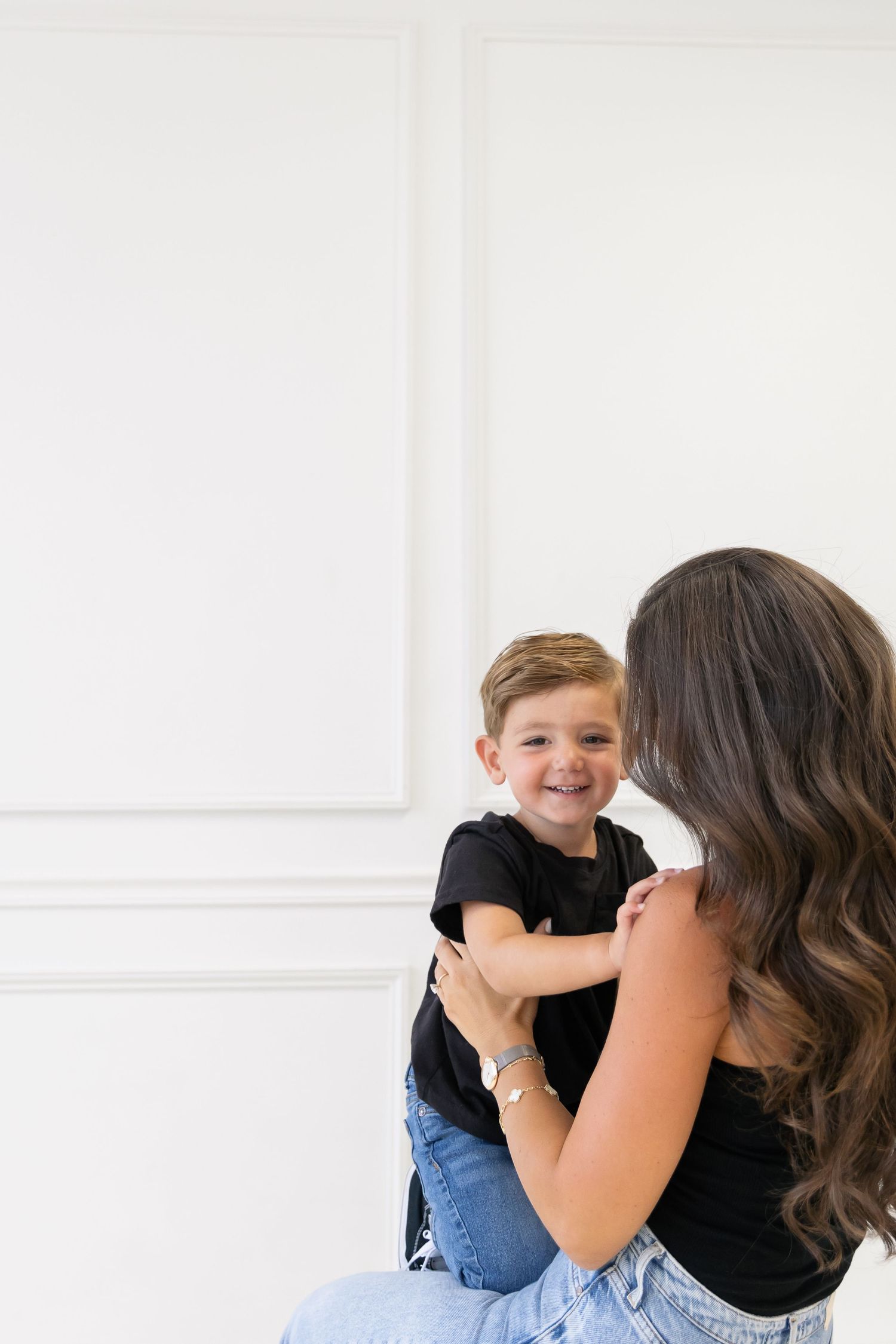A parent and child share a joyful moment together against a white wall background, both wearing casual attire.