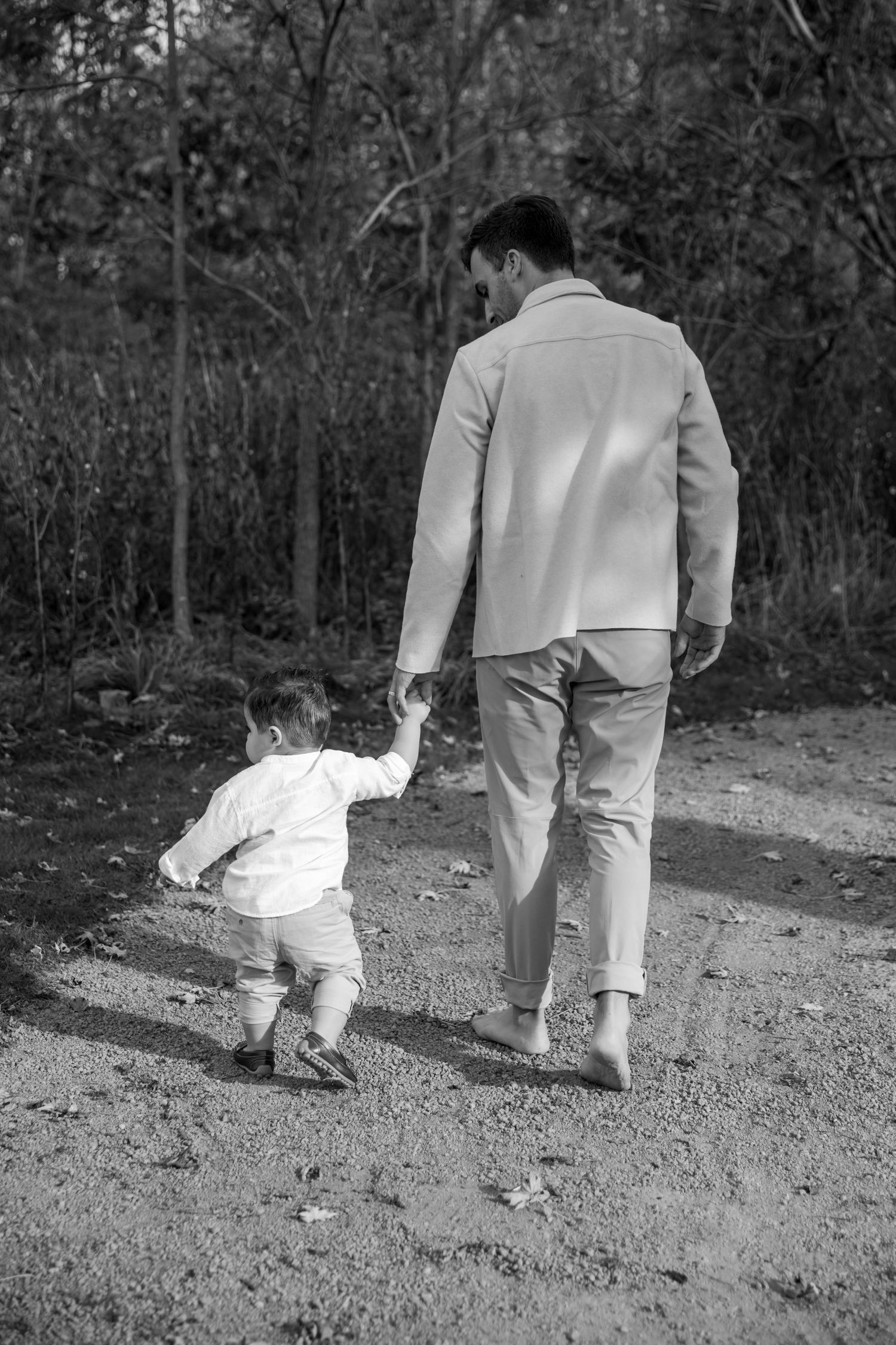Black and white photo of adult and small child walking hand in hand down a path in a wooded area.