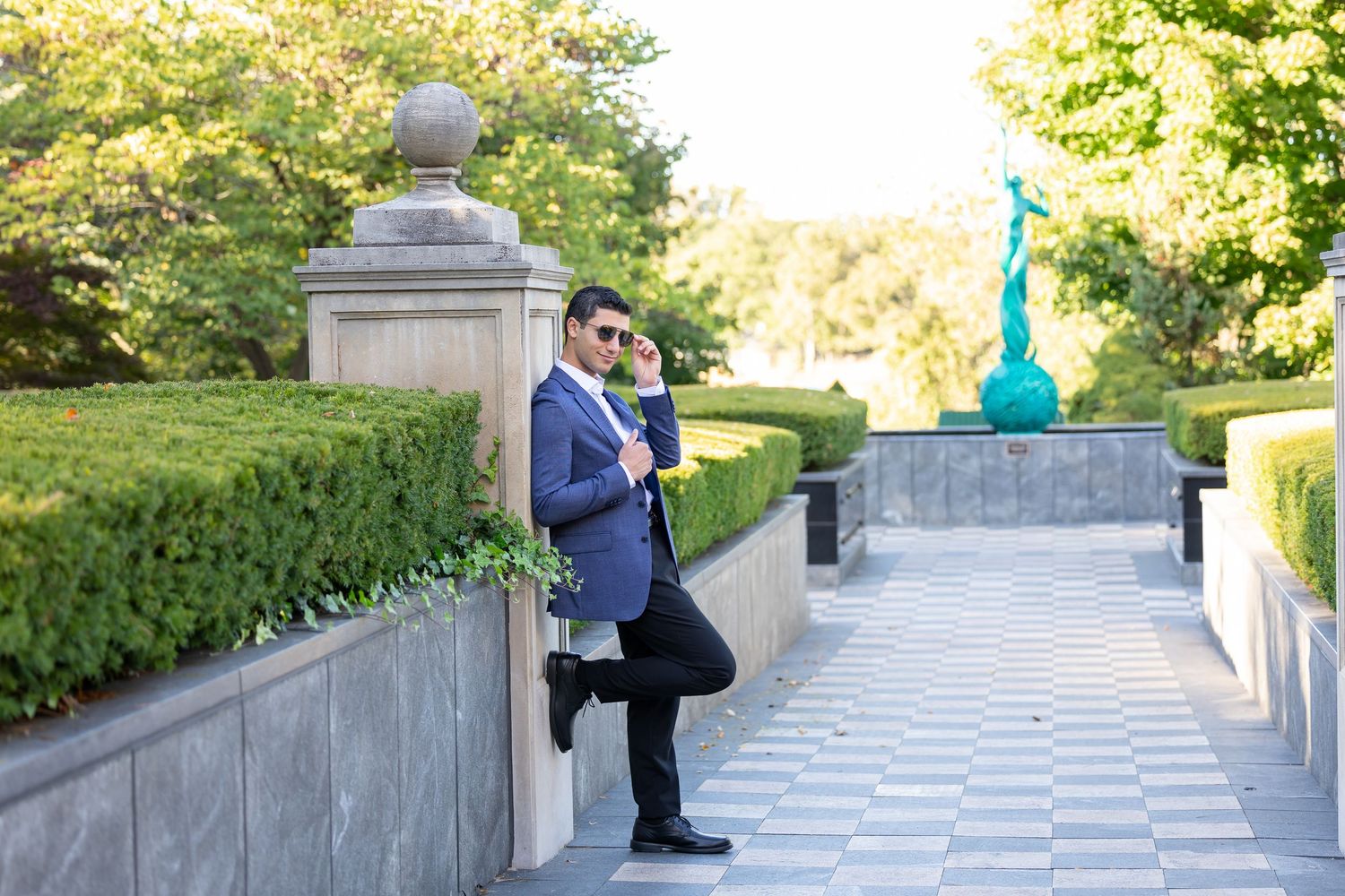 Person in formal attire leans against stone railing in manicured garden with decorative art.