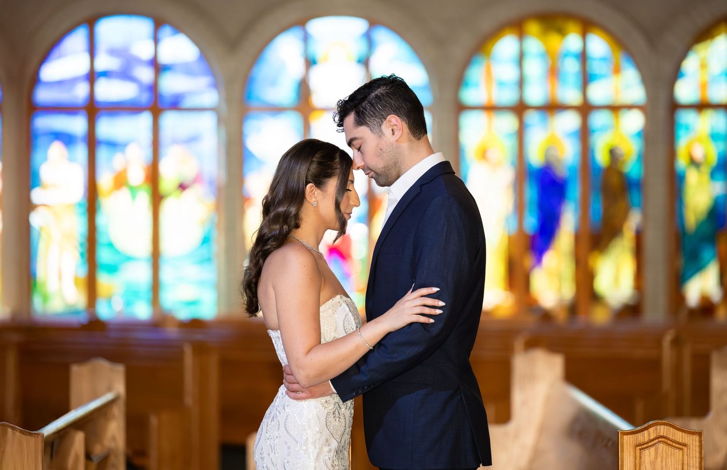 A couple in wedding attire stands intimately in front of stained glass windows in a church.