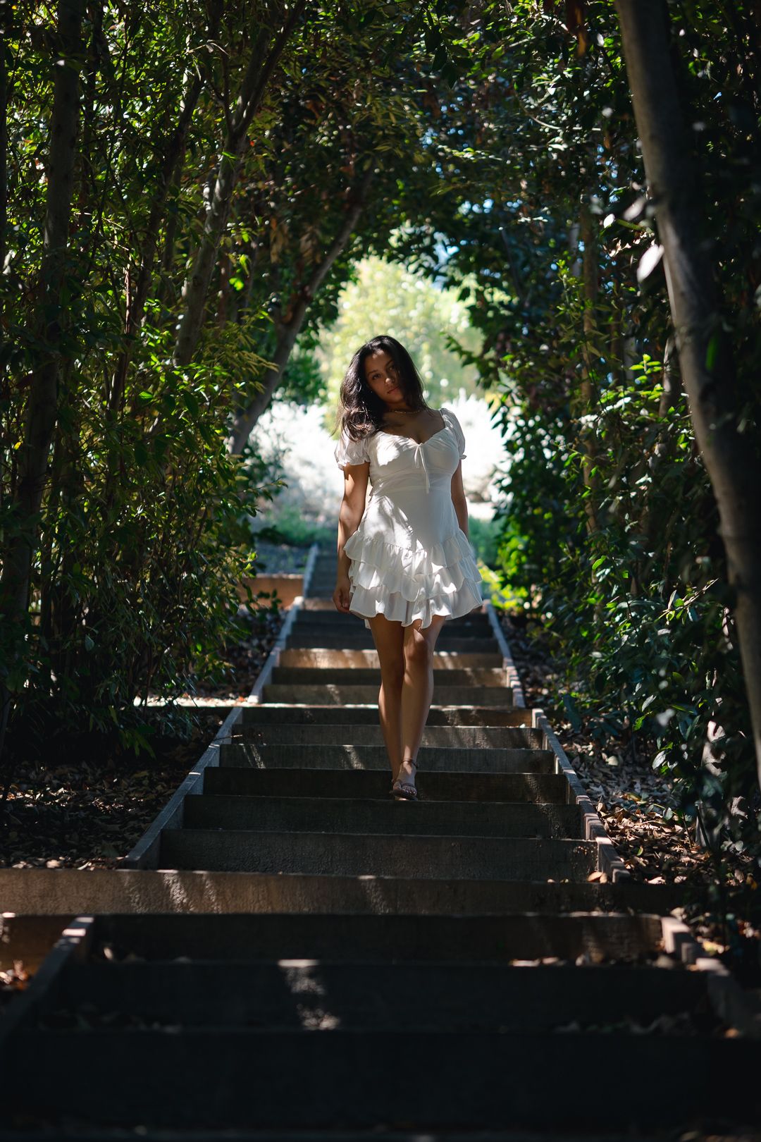 A person in a white dress descends stone steps through a lush green wooded archway on a sunny day.