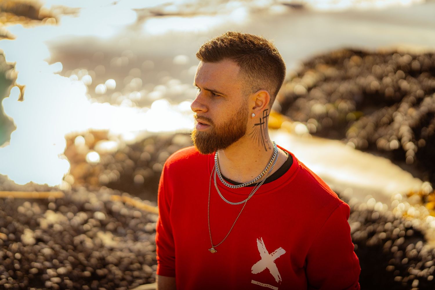 Person in red sweater with cross design poses against dramatic rocky coastal sunset landscape.