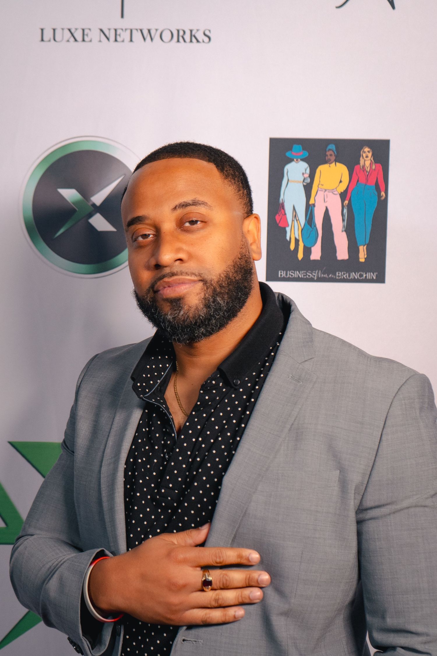 Professional portrait of man in grey suit and polka dot shirt against branded backdrop.