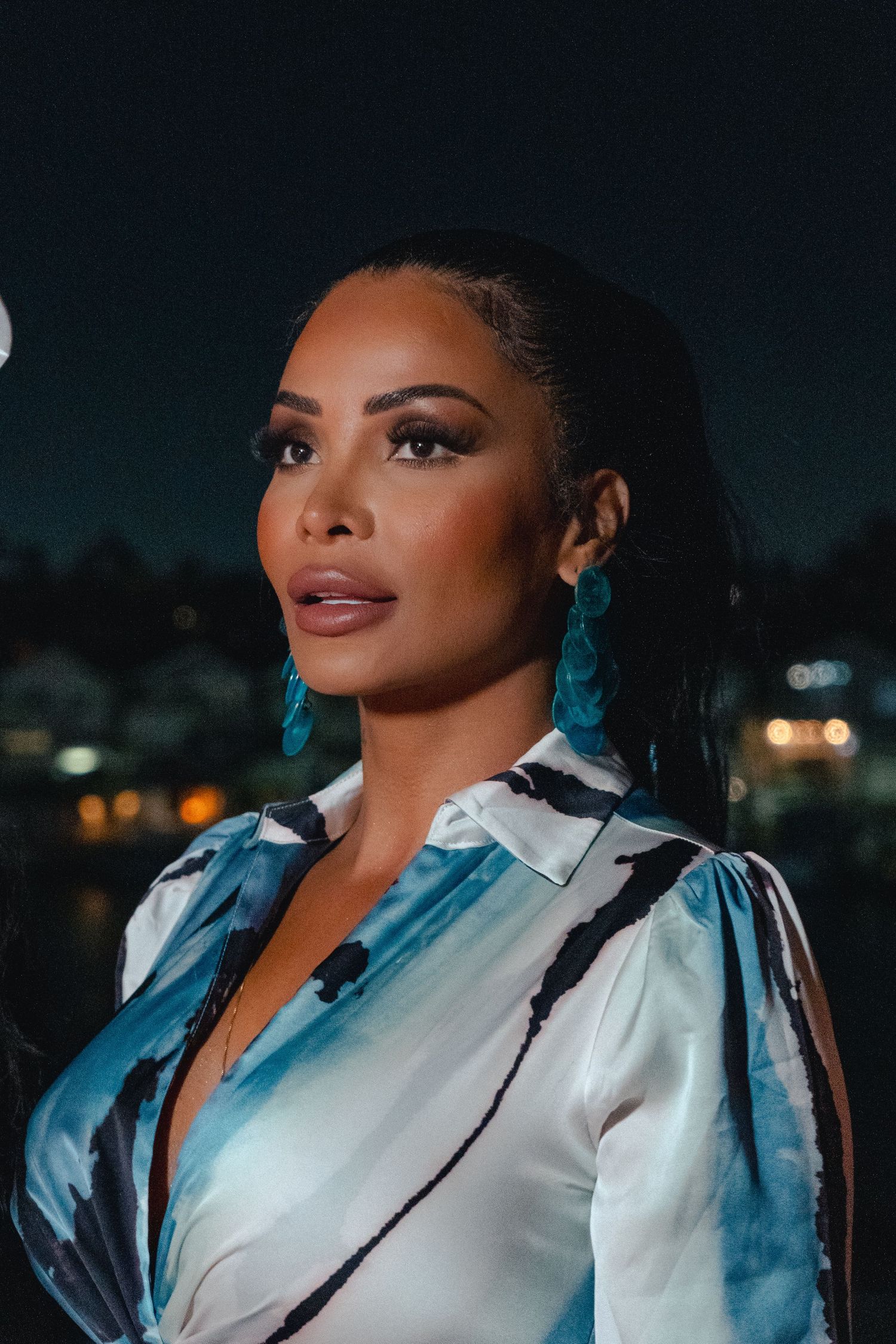 Portrait shot in evening light showing a person wearing turquoise earrings and a white patterned blouse against a dark cityscape.