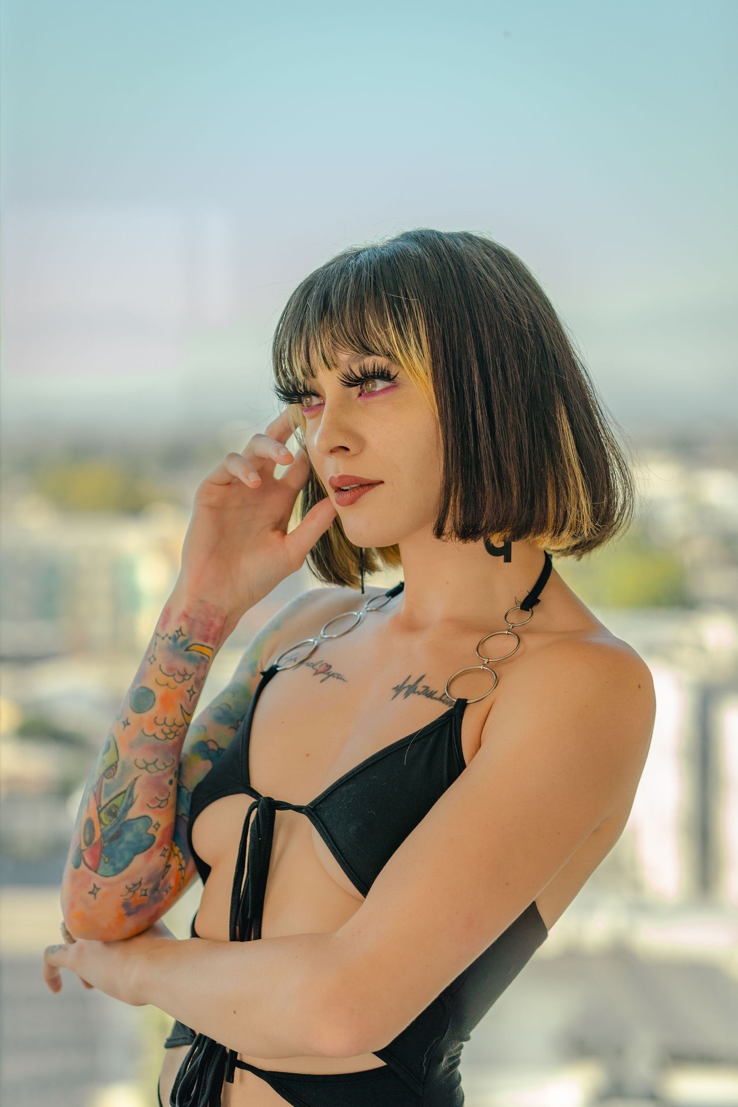 Tattooed woman in black bikini top poses thoughtfully on urban rooftop.