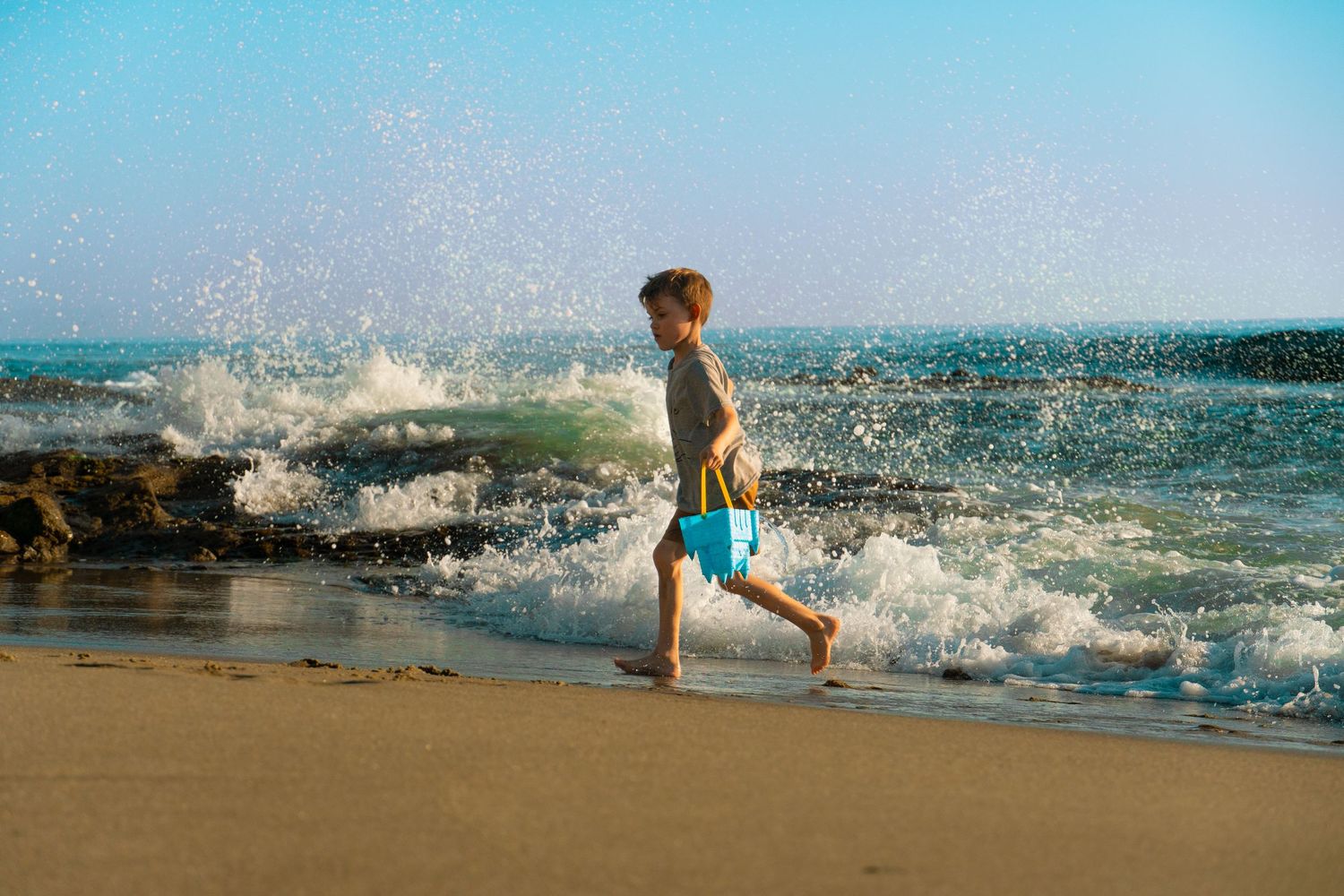 A person strolls along a sandy beach as ocean waves crash and spray water behind them on a bright sunny day.