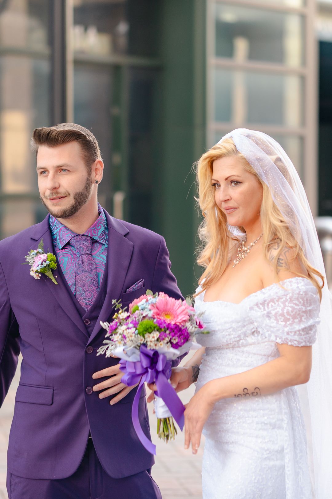 Wedding couple in purple suit and white dress holding purple floral bouquet.