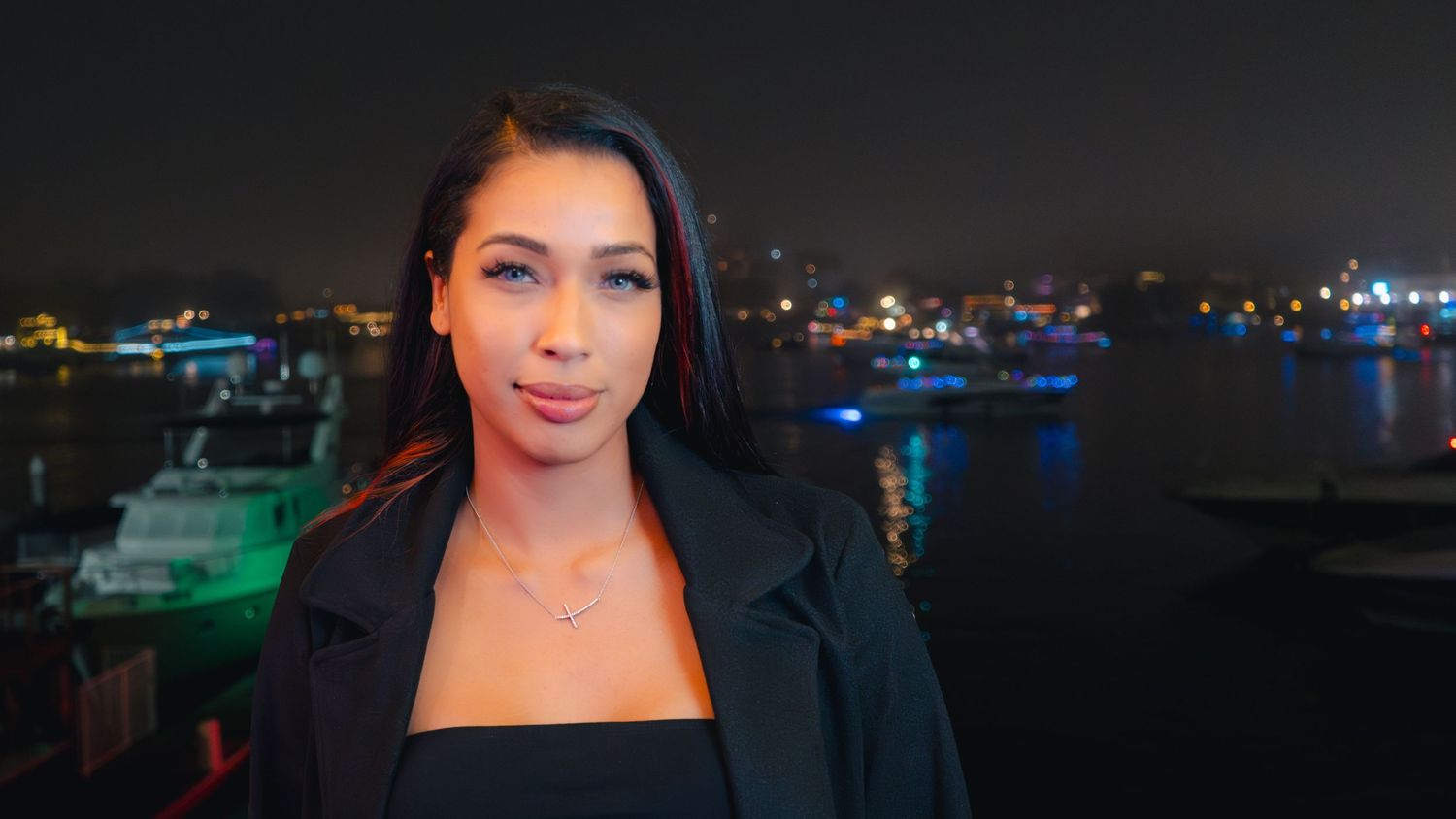 Evening portrait shot with boats and harbor lights visible in the background.